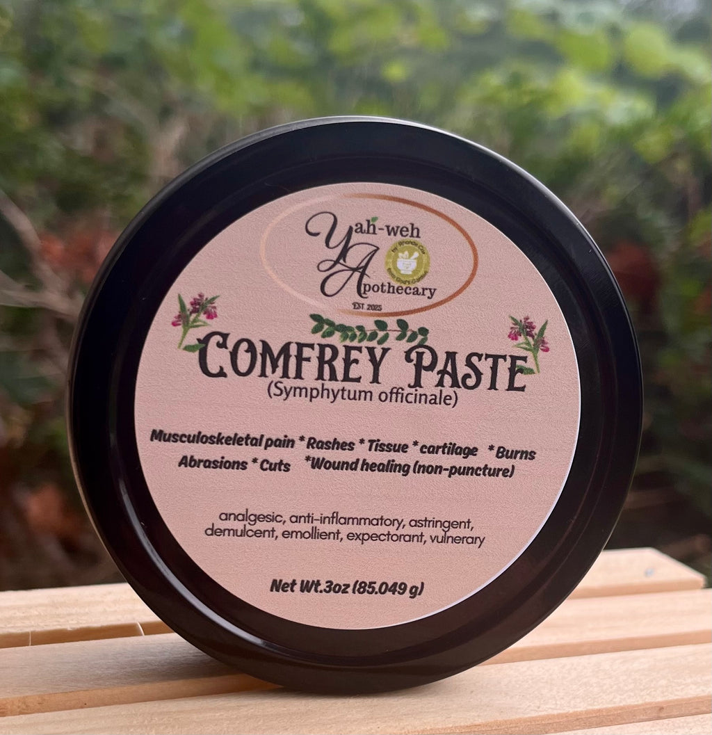 A round black plastic tub of Yahweh's Apothecary Comfrey Leaf Paste sits on a wooden surface with a blurred green outdoor background. The label is a light chocolate brown with black text and small botanical illustrations
