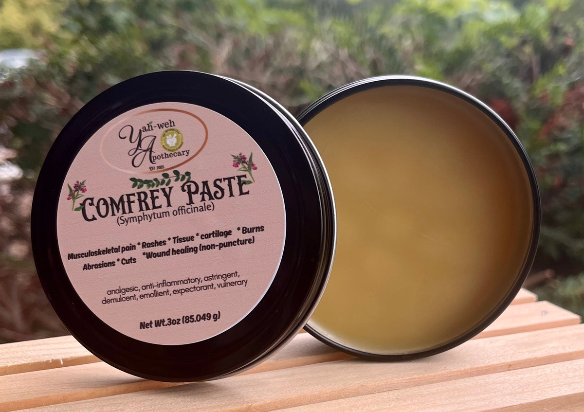 An open round metal tin of Yahweh's Apothecary Comfrey Leaf Paste sits beside its lid on a wooden surface with a blurred outdoor garden background.