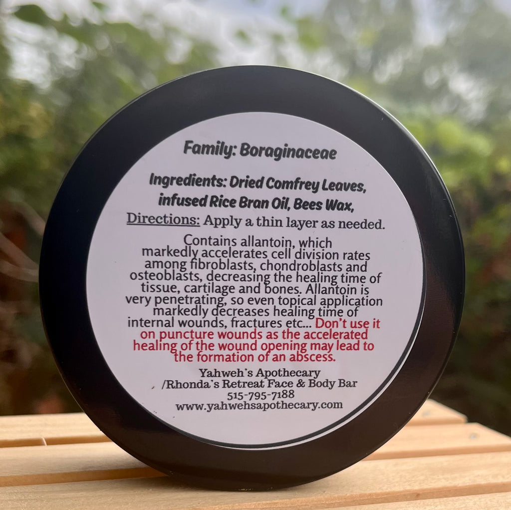 A close-up of the Comfrey Leaf Paste round black lid with a white printed label, photographed outdoors on a wooden surface with blurred greenery in the background. The label reads 'Family: Boraginaceae' and includes ingredients, directions, and contact information for Yahweh's Apothecary.