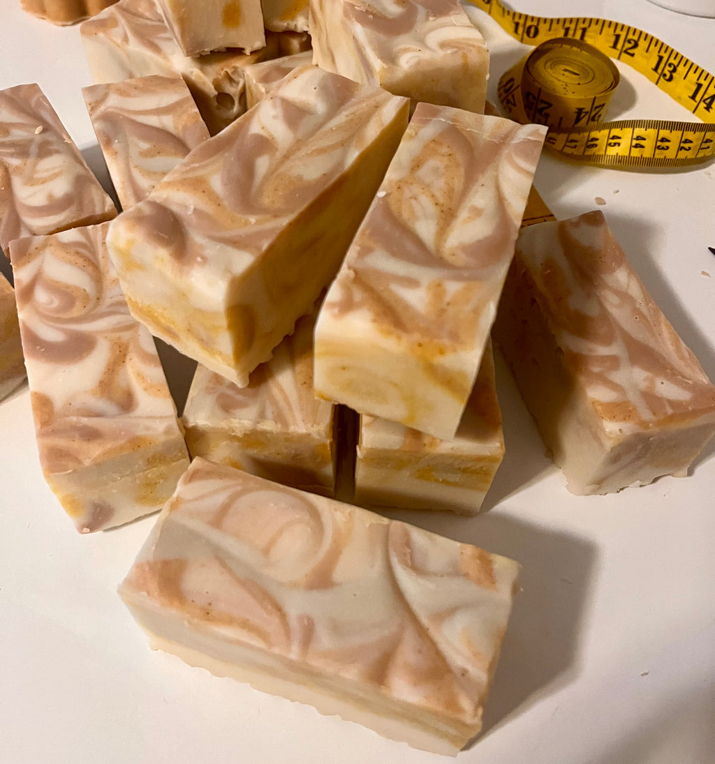 Several rectangular bars of marbled soap in warm cream, beige, and peach tones are arranged on a white surface, with a yellow measuring tape visible in the background.