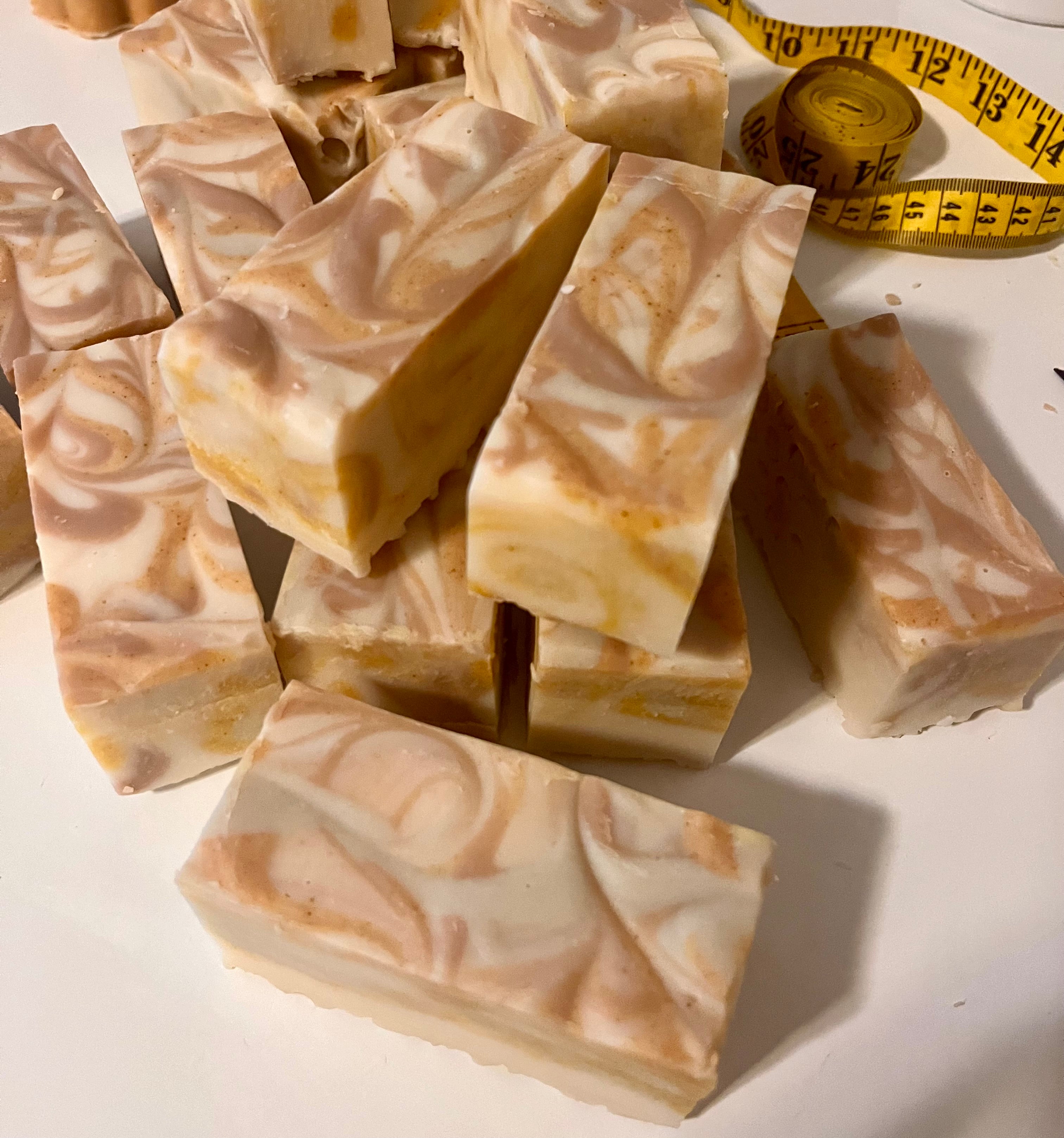 Several rectangular bars of marbled soap in warm cream, beige, and peach tones are arranged on a white surface, with a yellow measuring tape visible in the background.